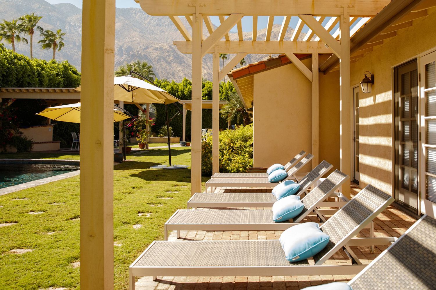 Row of wooden lounge chairs with blue pillows under a pergola beside a sunny pool in Palm Springs.