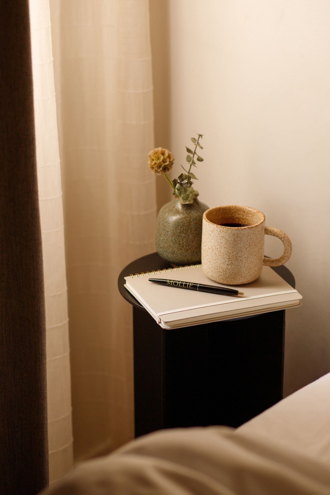 A cozy bedside table with a speckled ceramic mug, open notebook, pen, and small vase with dried flowers in warm light.