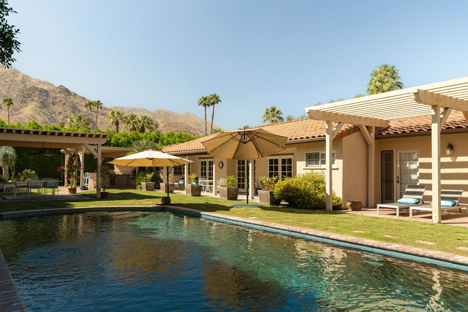 Luxury Palm Springs home with sparkling pool, pergola, patio umbrellas, and mountain views on a sunny day.