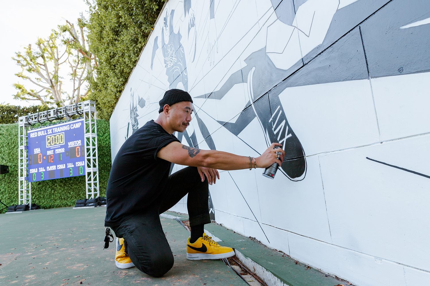 Artist works on a large-scale mural painting with geometric shapes in white and gray tones on an outdoor wall.