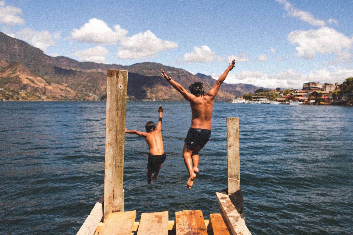 Two people jumping off a wooden dock into a scenic mountain lake on a sunny summer day.