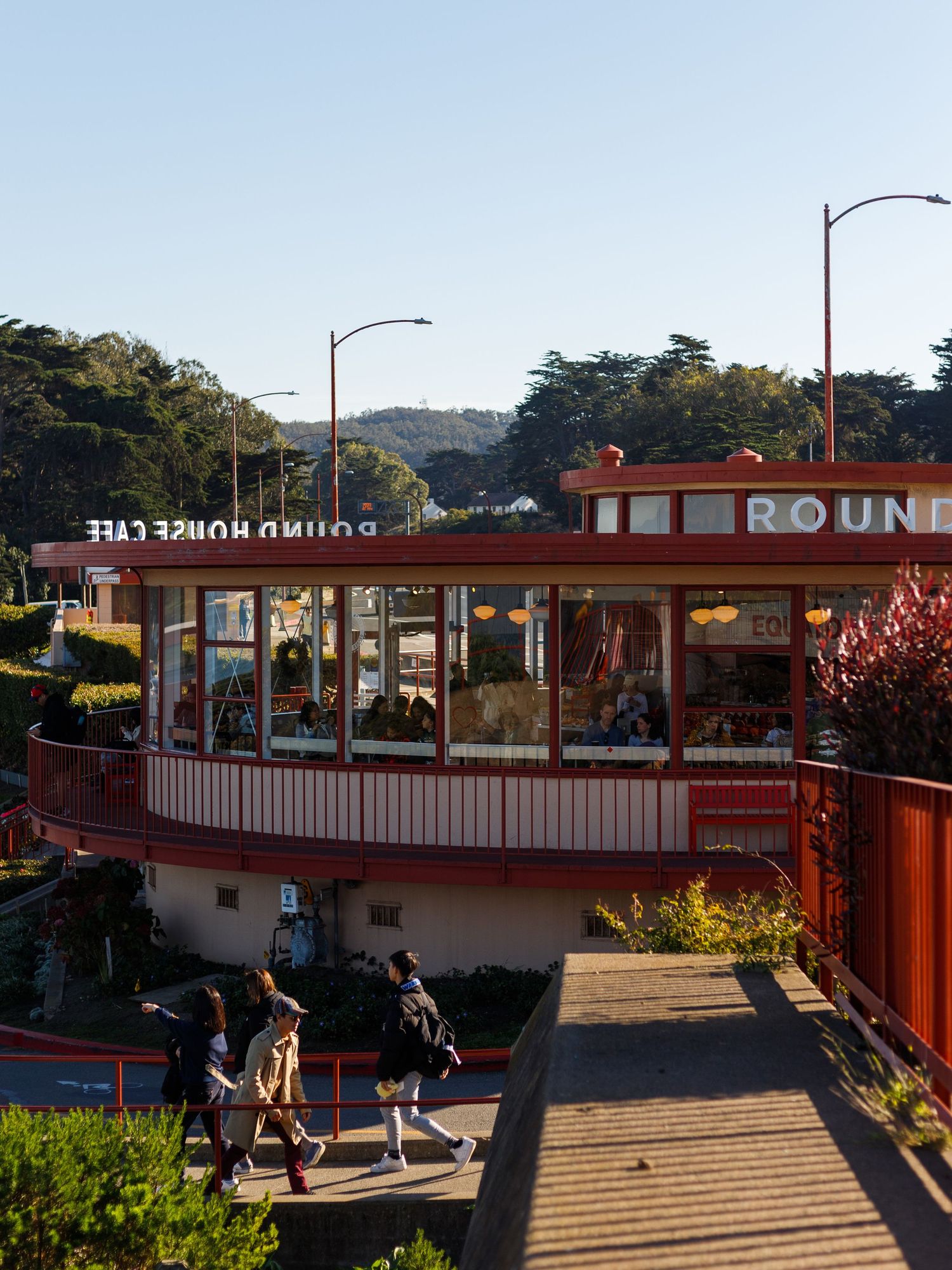 People walk past the Round House Cafe, a red circular building in San Francisco's Golden Gate Park on a sunny day.