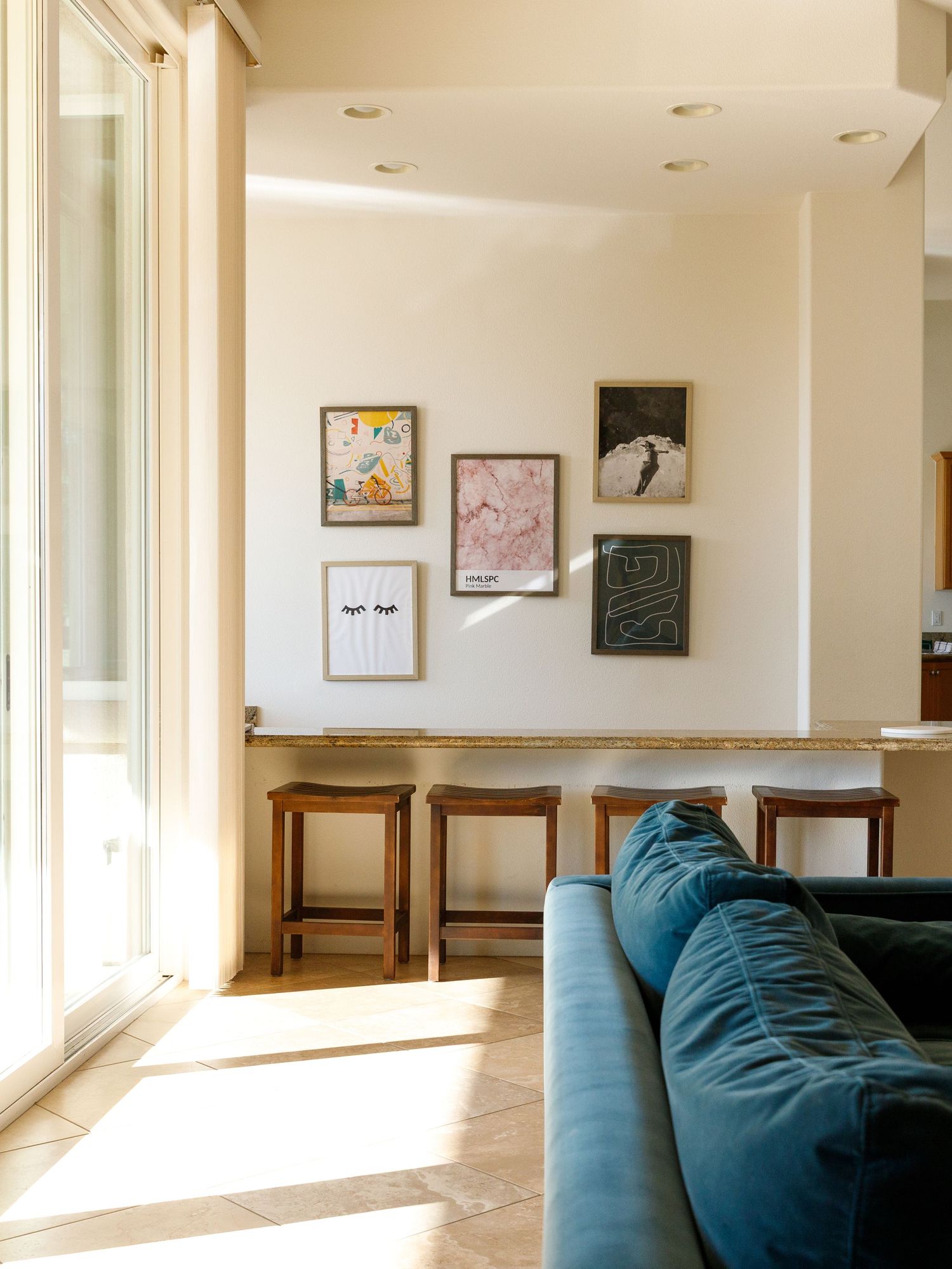 Bright living room with teal velvet sofa, gallery wall art, wooden bar stools, and sunlit herringbone tile floors.
