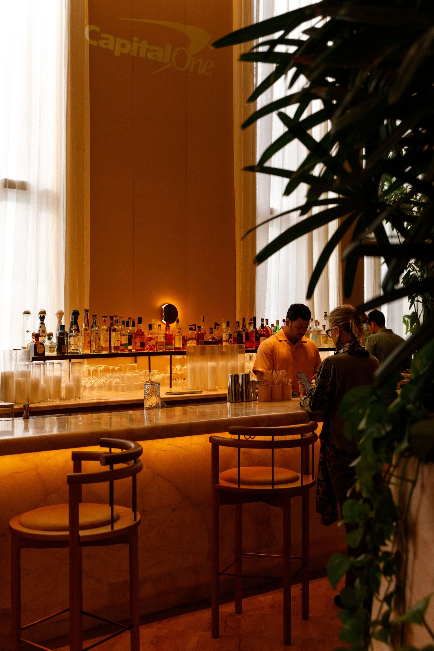 Warm Capital One bar lounge with bartender, marble counter, backlit liquor shelves, bar stools, and tropical plants.