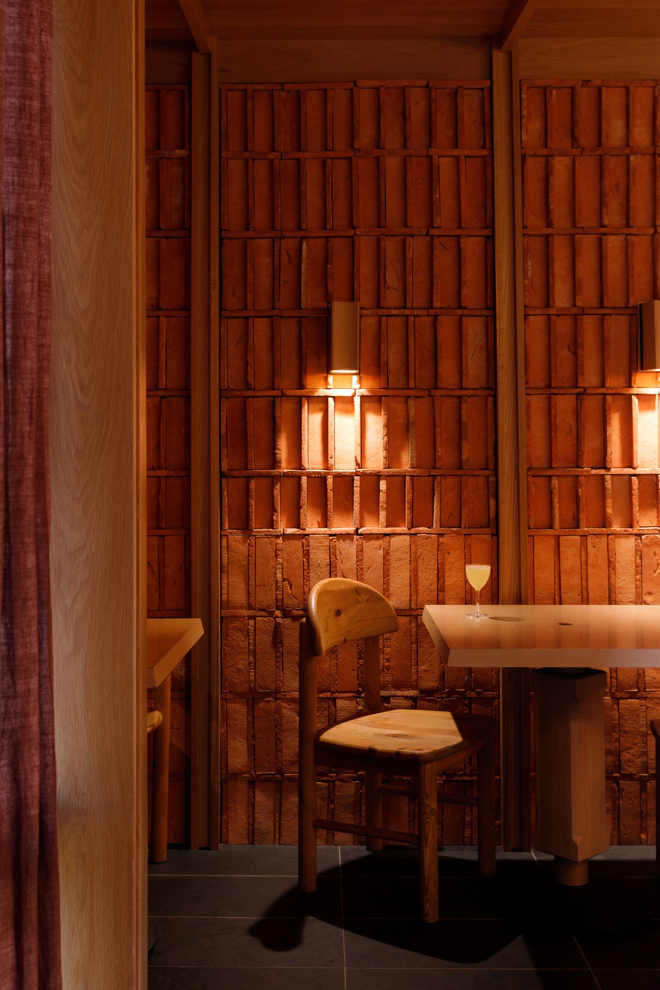 Cozy restaurant booth with warm amber lighting, wooden walls, a light wood table and chair, and a wine glass on the table.