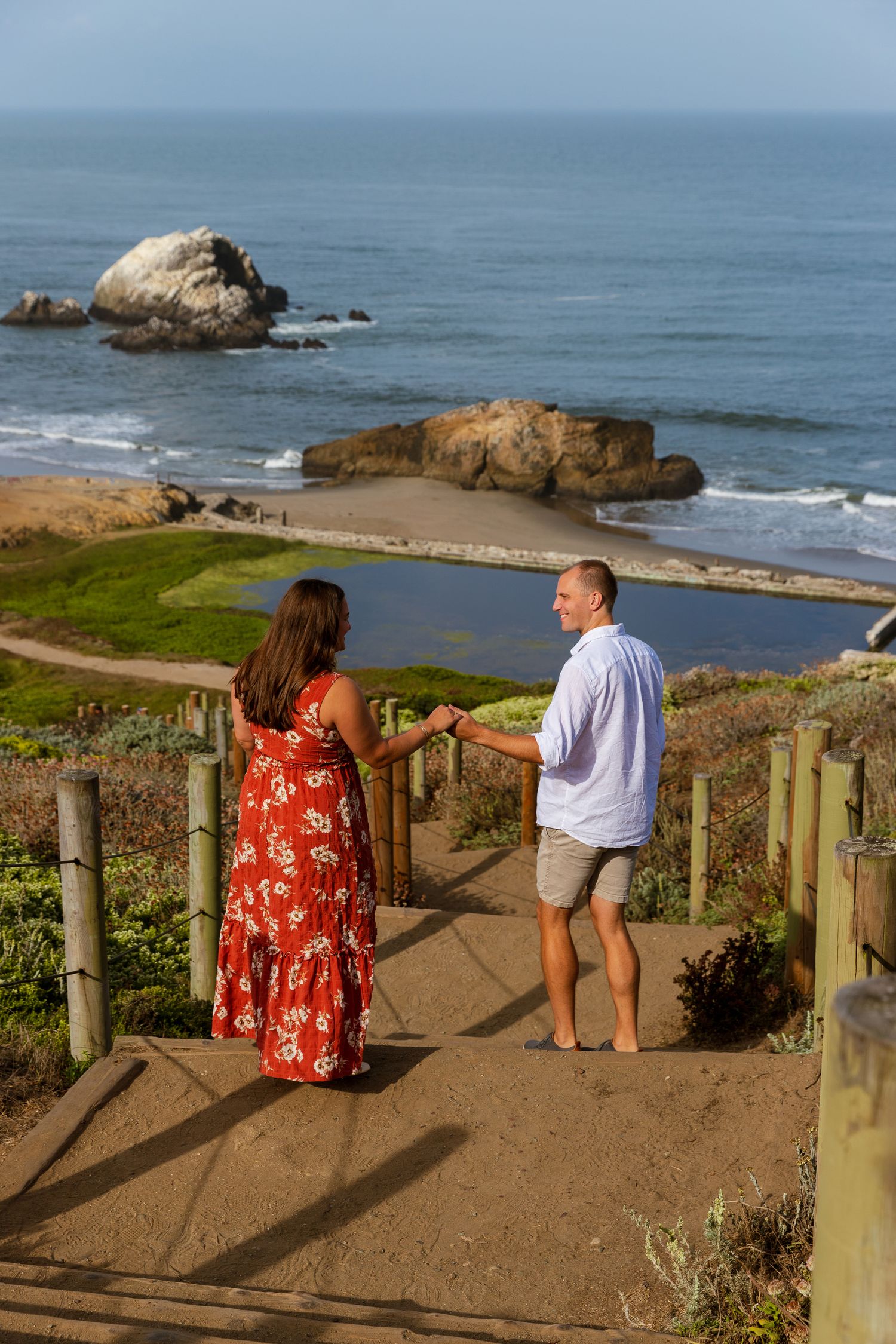 Two people stand on a coastal path overlooking a scenic beach with large rock formations and ocean waves in San Francisco.