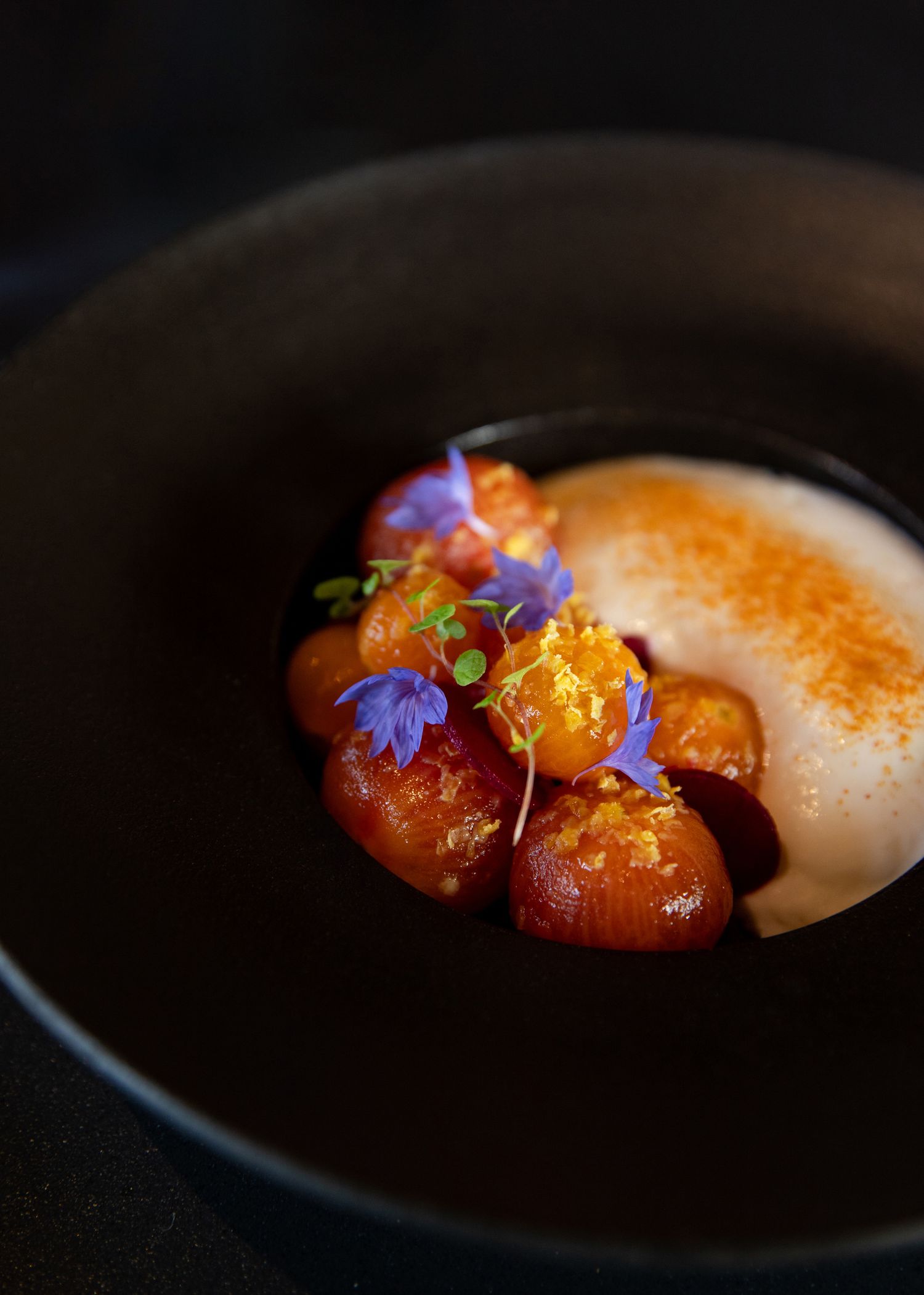Elegant plated dessert with colorful edible flowers and sauce on a black ceramic dish. Food photography for luxury restaurants and hotels.