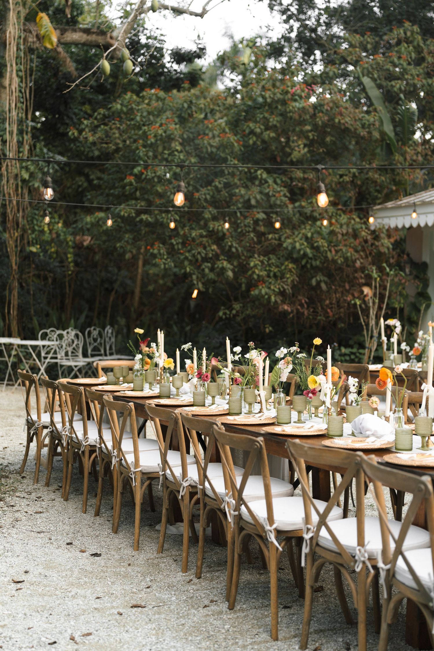 Long rustic wooden tables with white linens set for outdoor dining under string lights and tree canopy at a garden event.
