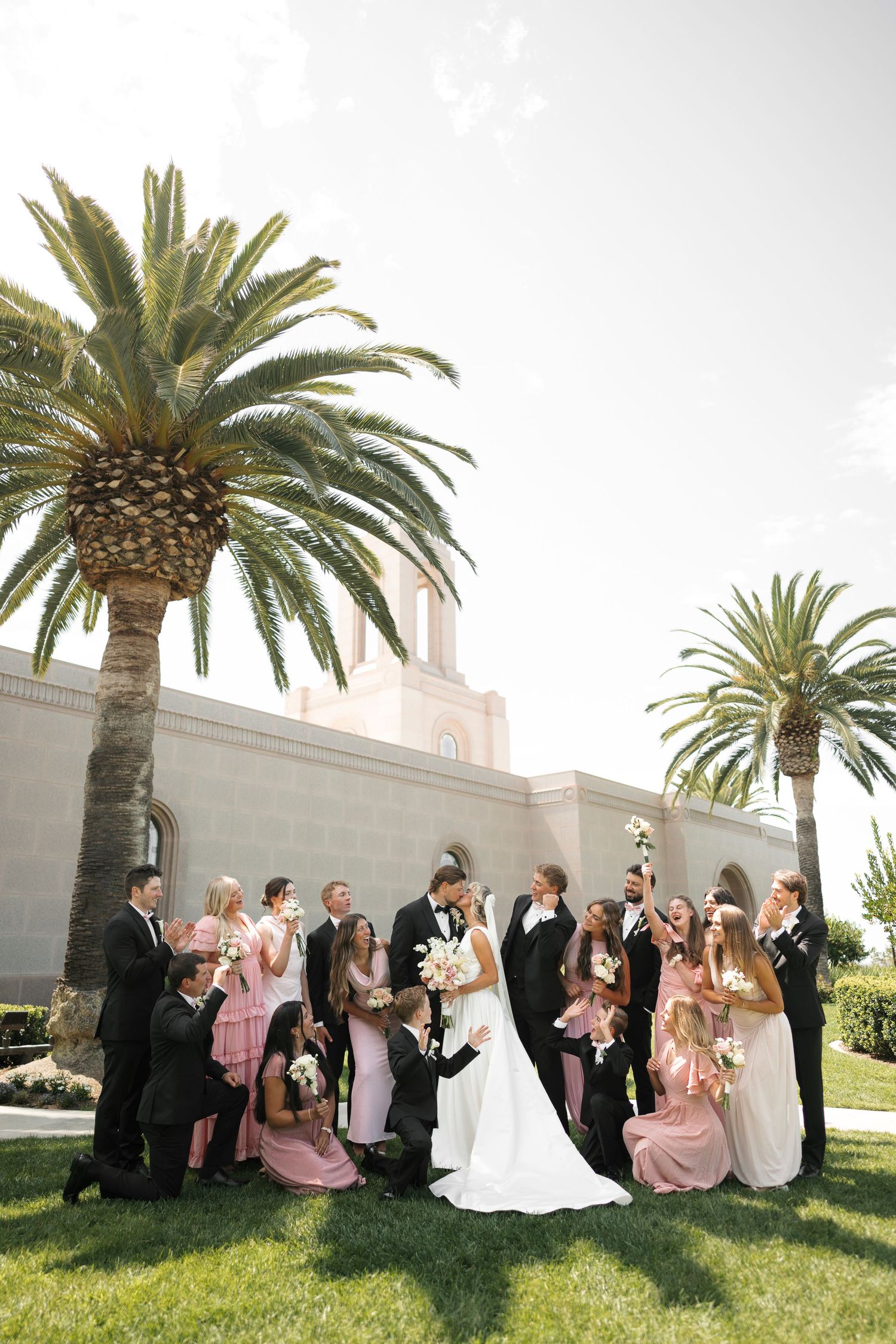 Wedding party poses with bride and groom before a white building flanked by tall palm trees on a sunny day.