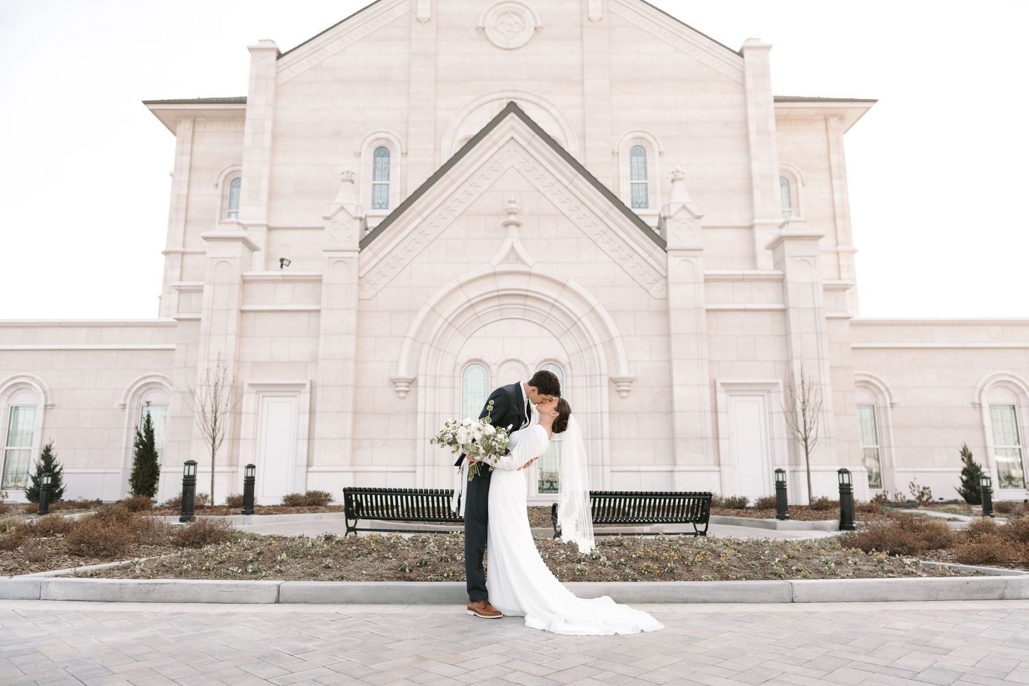 A couple in wedding attire shares a romantic moment outside a grand white church building.