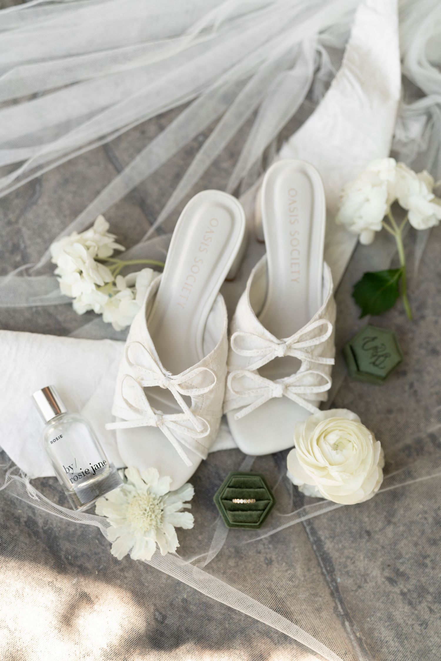 White bridal heels with bow detail beside perfume, ring box, and white flowers on a veil.