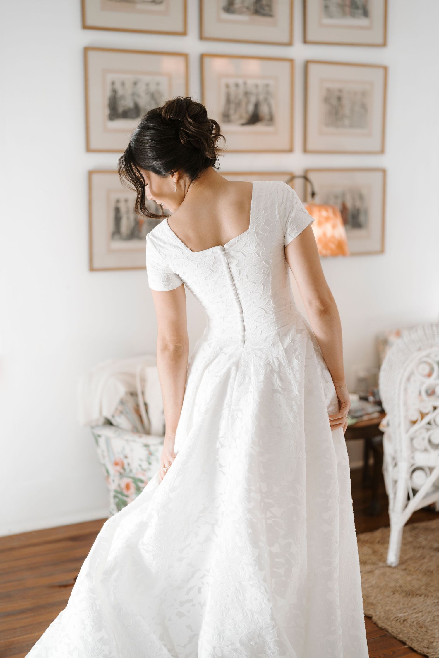 A white lace dress is shown in multiple angles on a figure against a wall with framed artwork.
