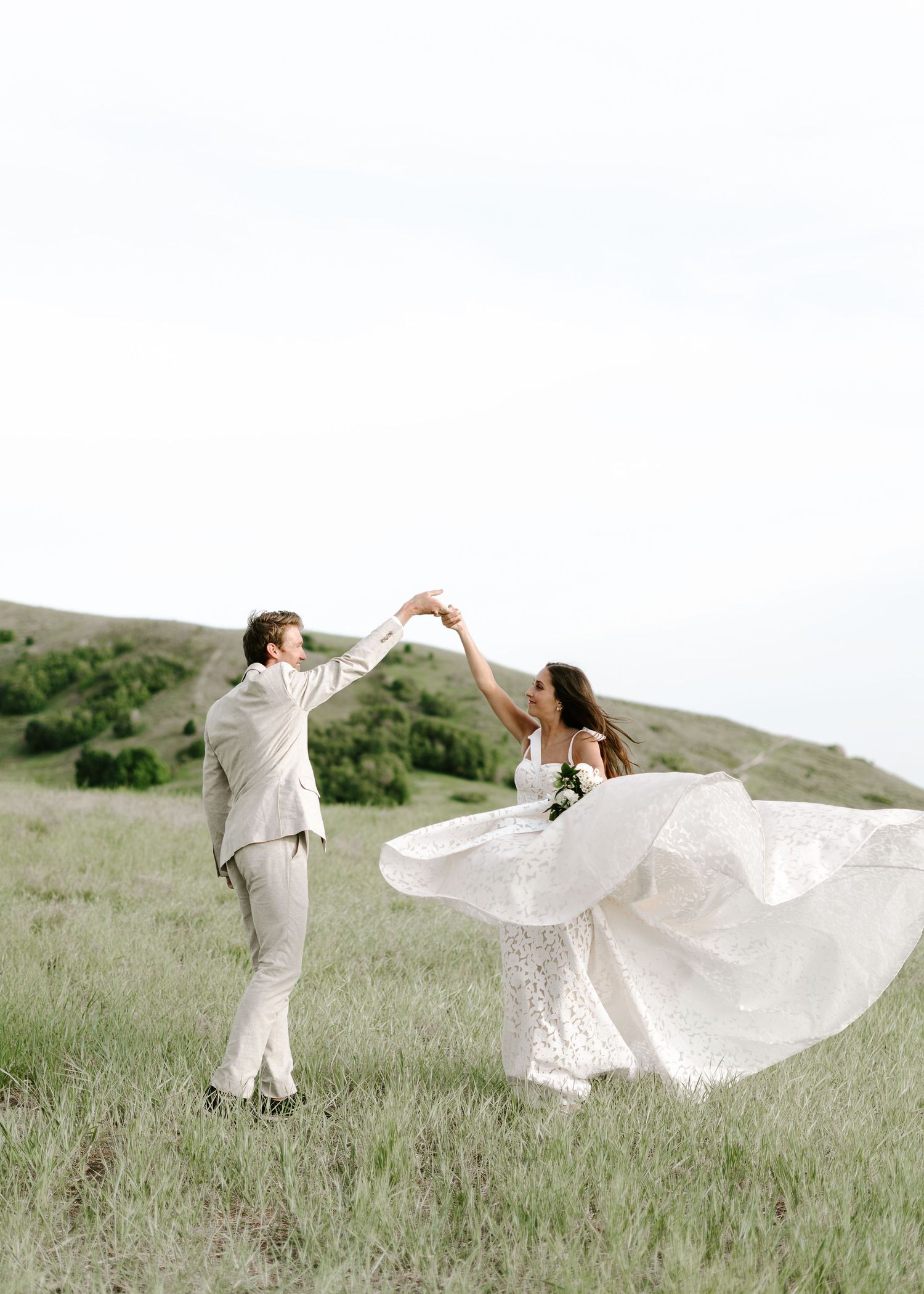 A couple in white outfits dances gracefully across a grassy hillside with flowing fabric creating ethereal movement.
