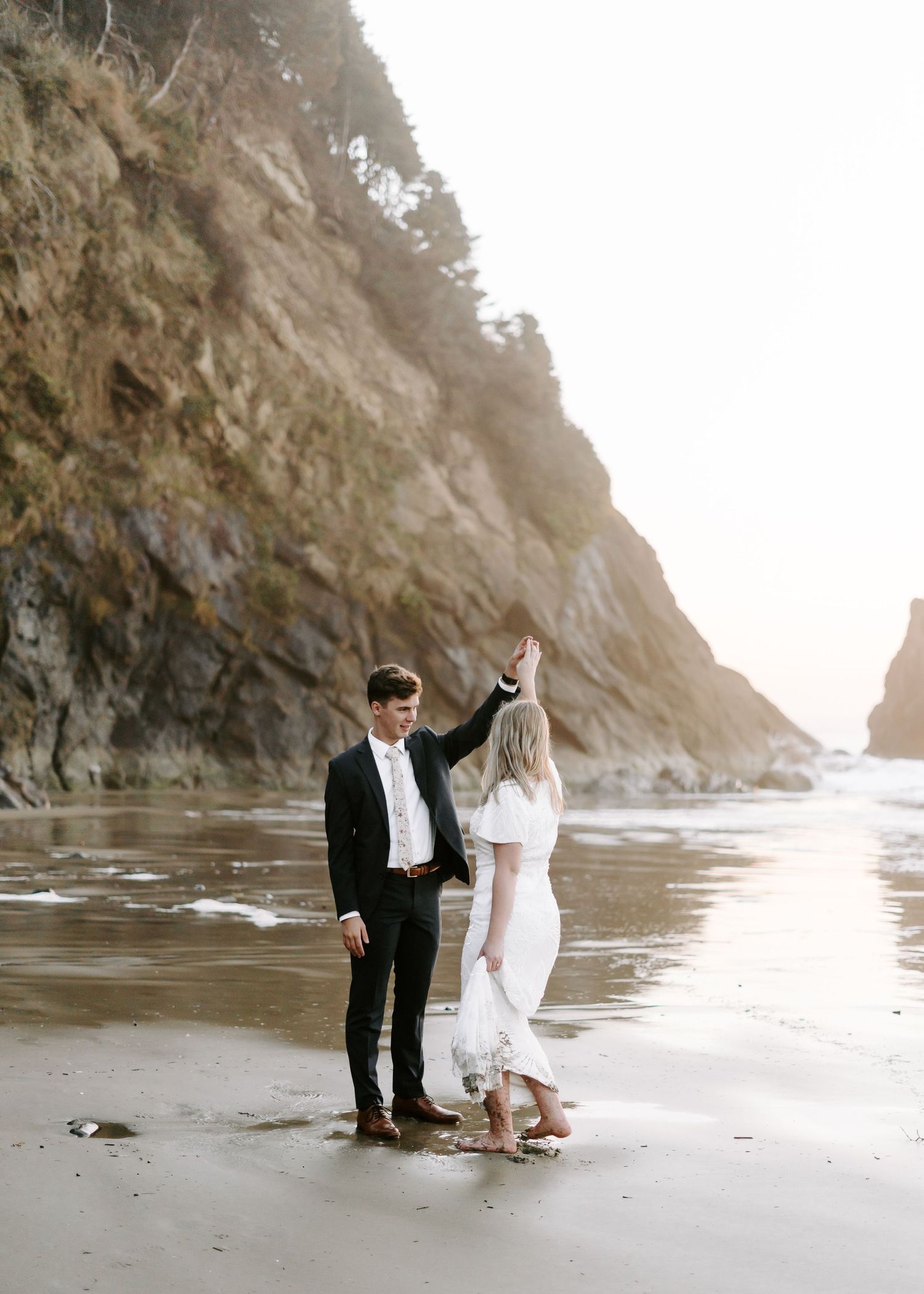 A wedding couple poses on a dramatic beach with towering cliffs and misty oceanside backdrop.