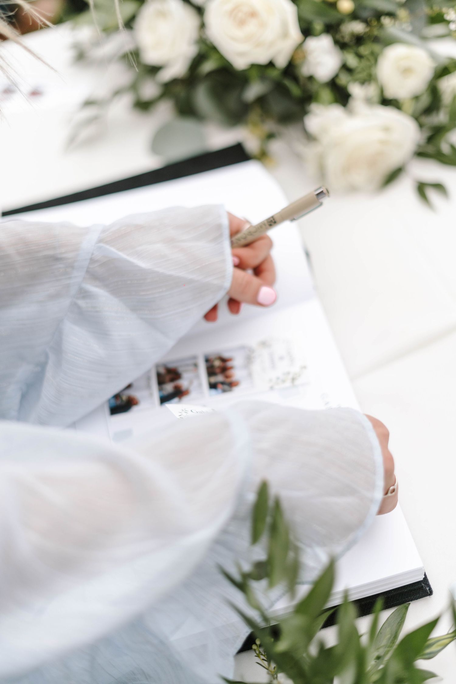 Person in white clothing writing in a guestbook surrounded by white roses and greenery.