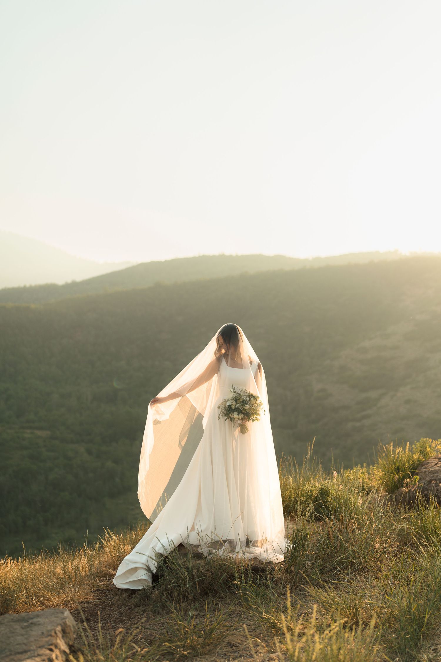 Bride in white gown and flowing veil stands on mountain cliff holding bouquet at golden sunset.