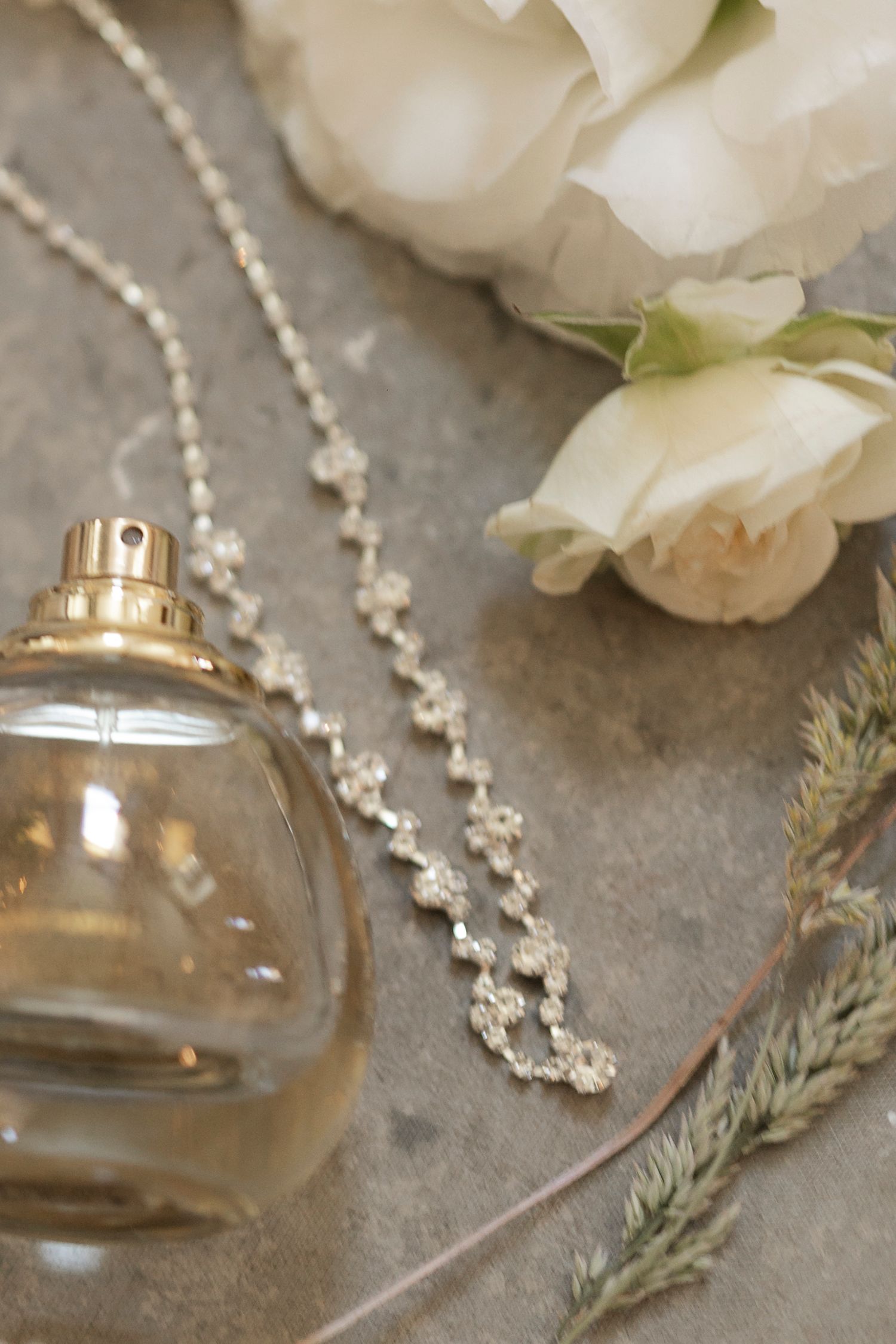 Elegant bridal flat lay with gold perfume bottle, crystal chain necklace, white roses, and dried botanicals on stone.