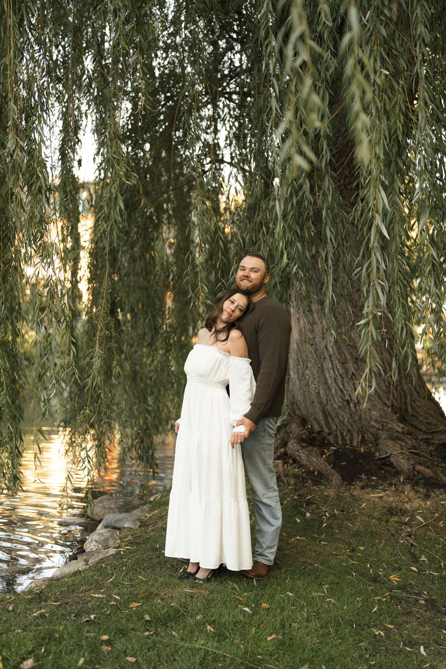 A couple embraces under a weeping willow tree beside a pond at sunset, with the woman wearing a white dress.