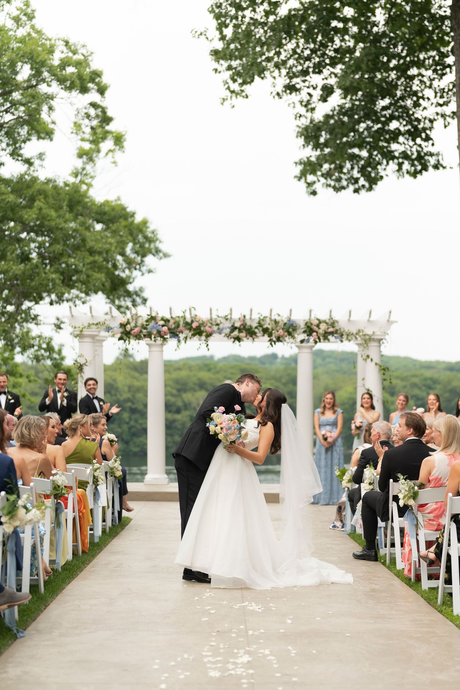 Bride and groom share first kiss at outdoor wedding ceremony with floral arch, white columns, and guests seated along aisle.