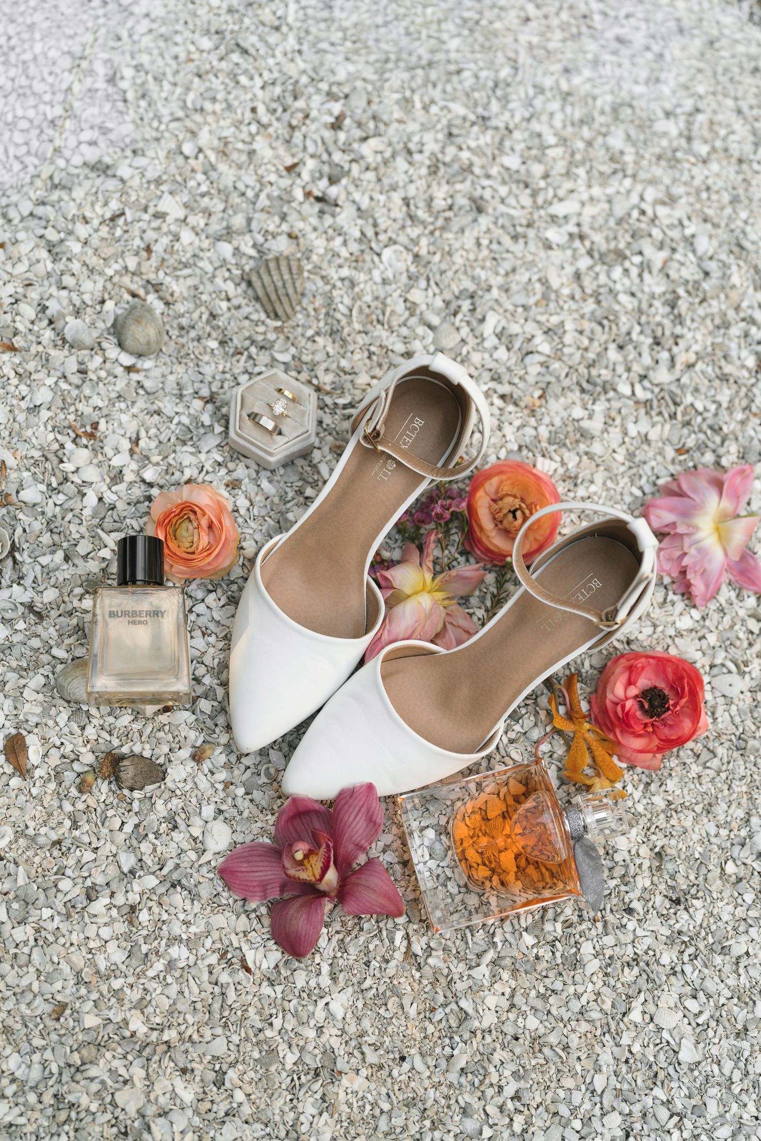 White bridal flats arranged with perfume bottle and colorful flowers including roses and orchids on textured concrete surface.