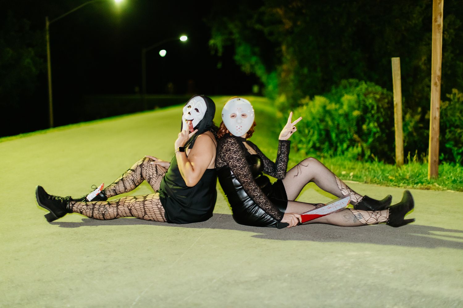 Masked individuals in dark clothing lying on illuminated pavement at night.
