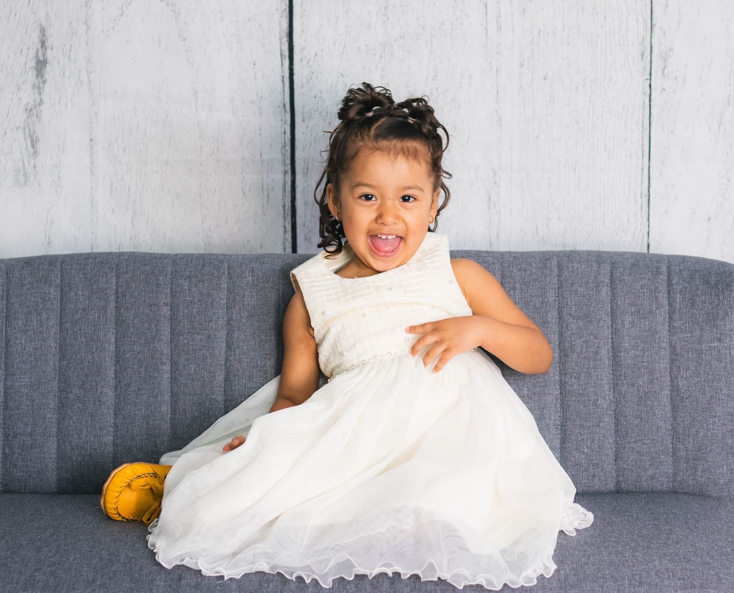 Series of playful poses of child in white dress on grey couch.