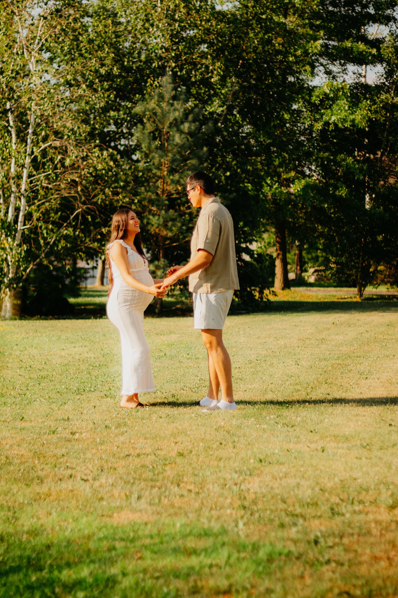 A couple in white outfits dances playfully in a sunlit grassy field during a summer evening.