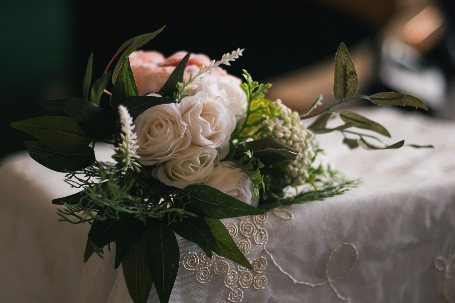 Elegant white roses and greenery wedding bouquet resting on lace-trimmed table at reception.