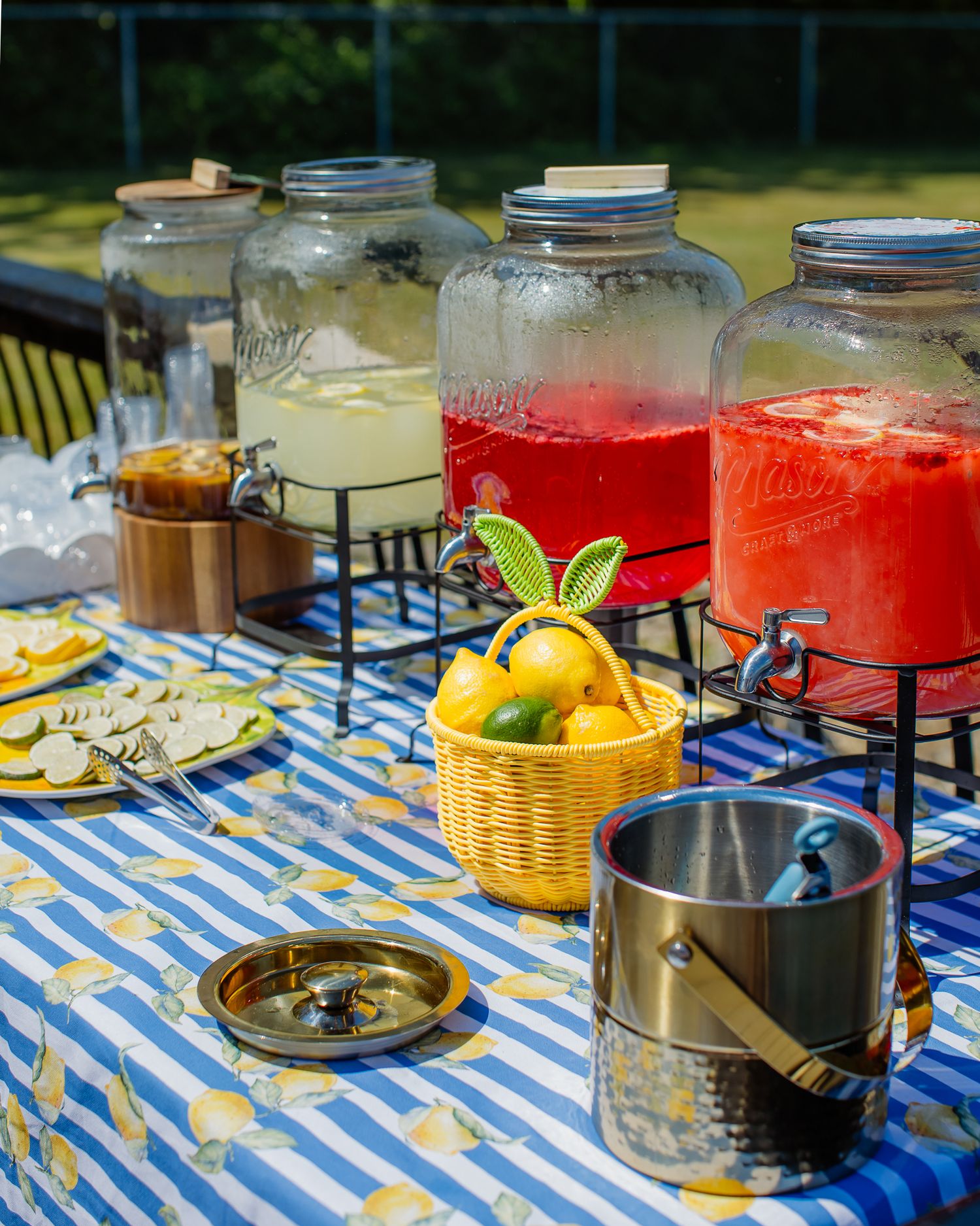 Outdoor party drink station with large glass dispensers of lemonade and punch on blue striped tablecloth.
