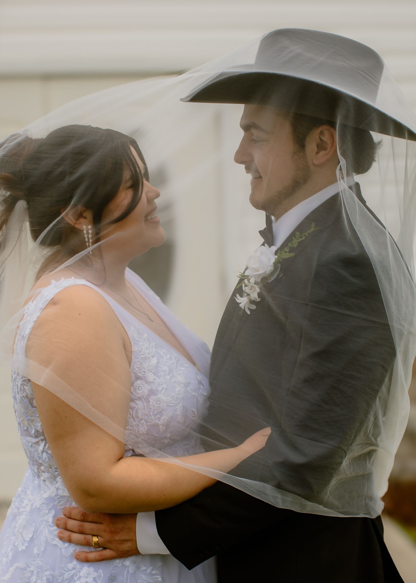 Bride and groom share a joyful moment under flowing veil, groom wearing a wide-brim cowboy hat.