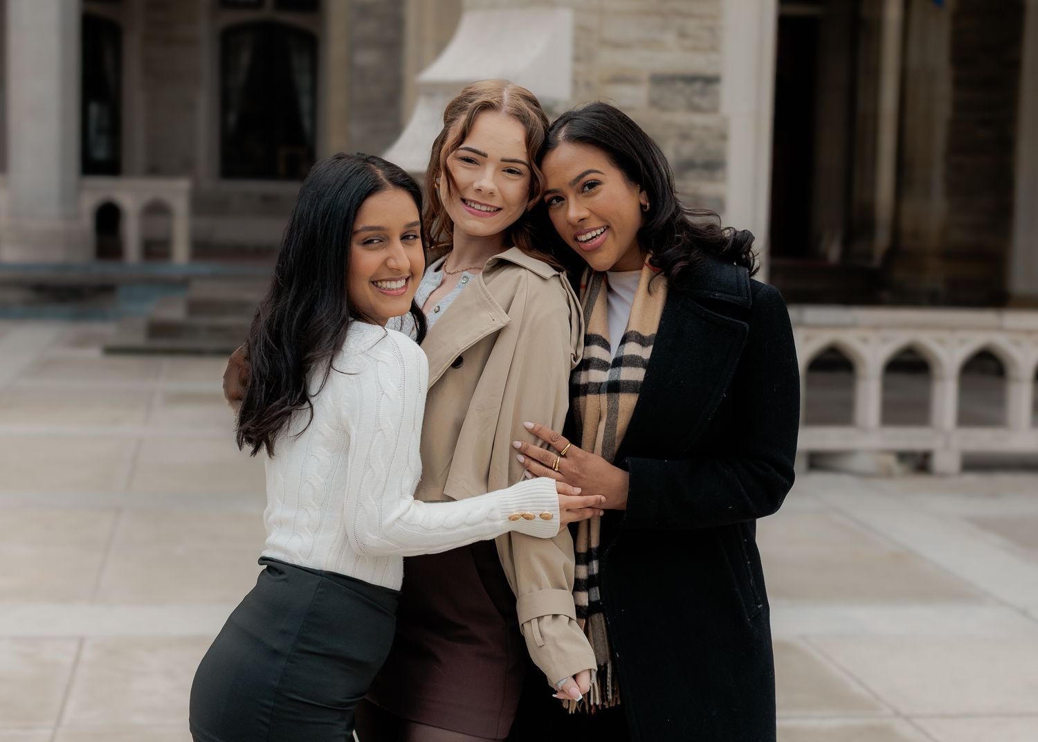 Three smiling women hugging outdoors near a stone building, wearing stylish fall outfits.