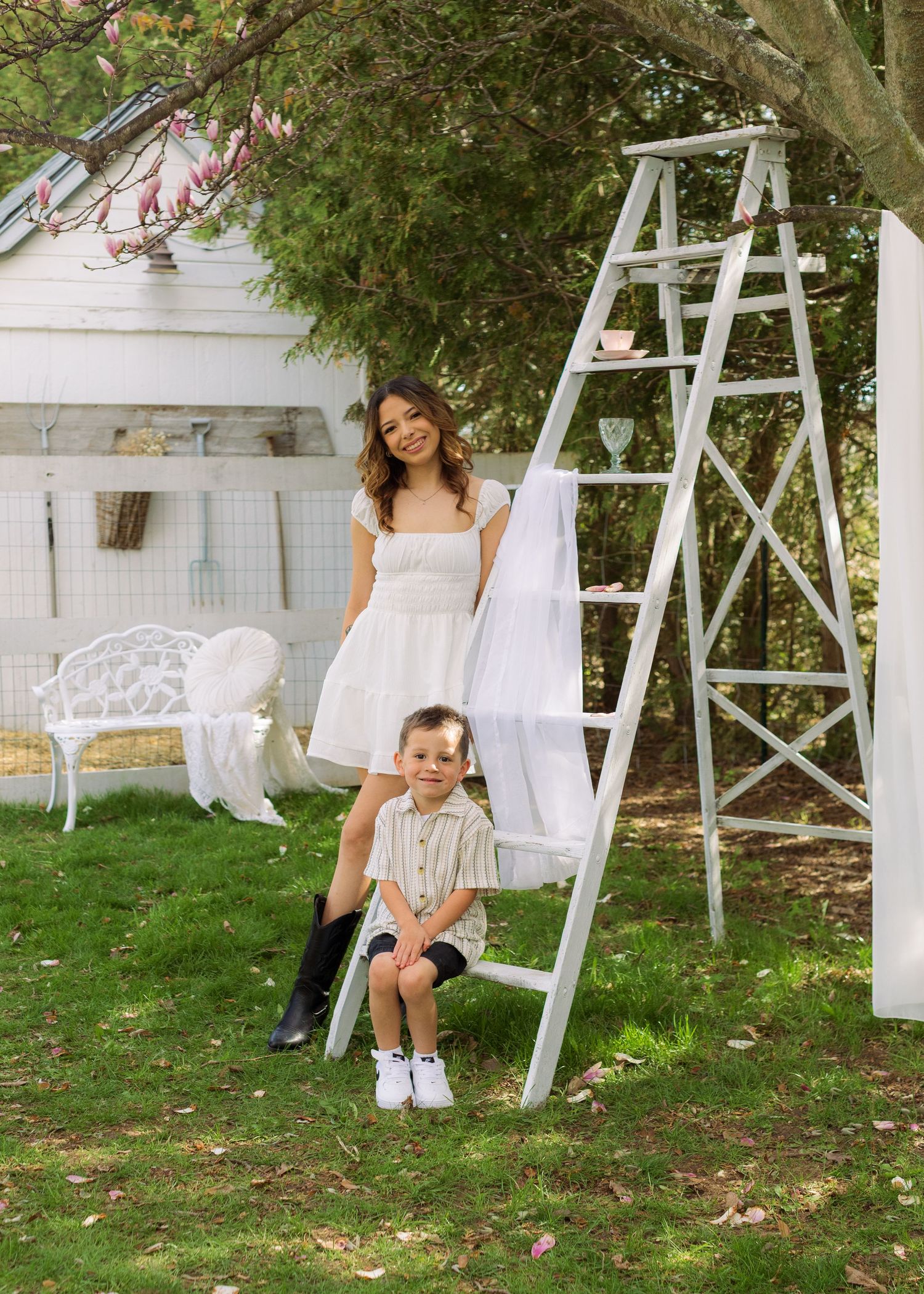 A woman in a white dress and boy in striped shirt pose by a decorative ladder swing in a lush garden.