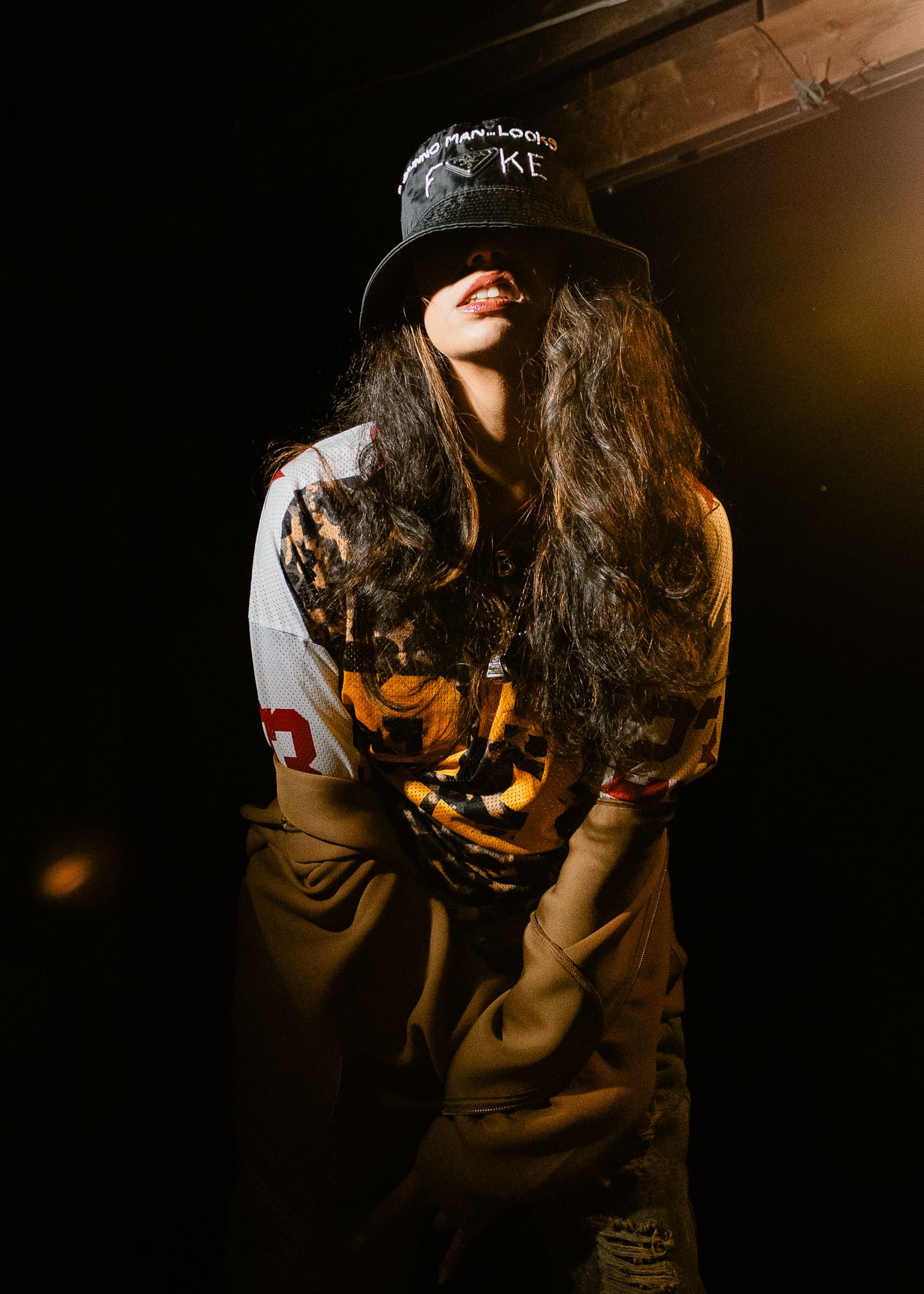Woman in bucket hat and graphic tee posing dramatically under dark moody lighting indoors.