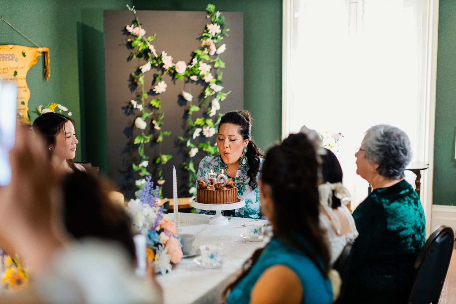 Woman blows out candles on a birthday cake surrounded by guests at an elegant indoor celebration.