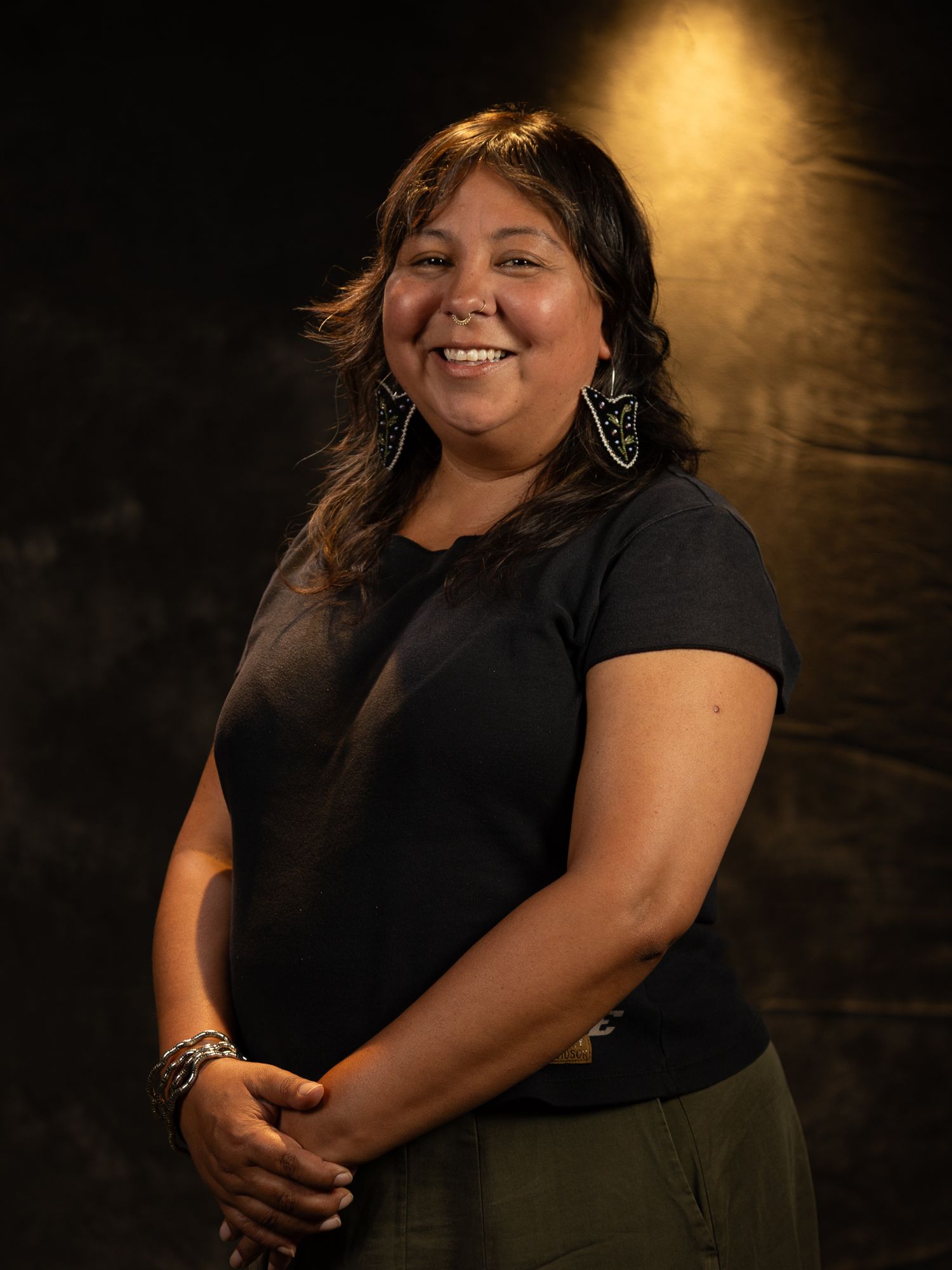 Smiling young woman with long dark hair wearing a black t-shirt and decorative earrings, posed against a dark background.