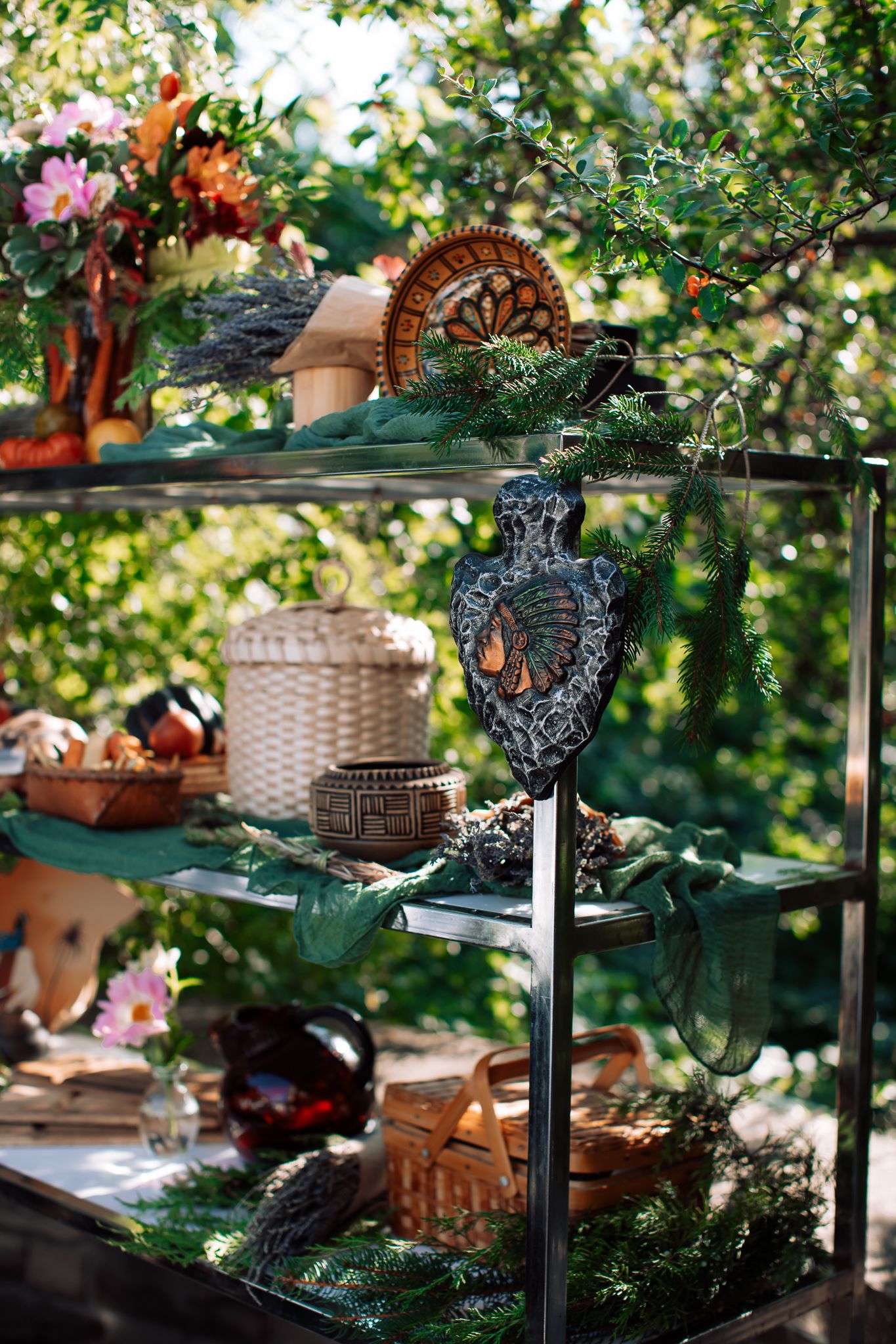 Outdoor display shelf with decorative items, wicker baskets, pine branches, and ornate crafts in a lush garden setting.