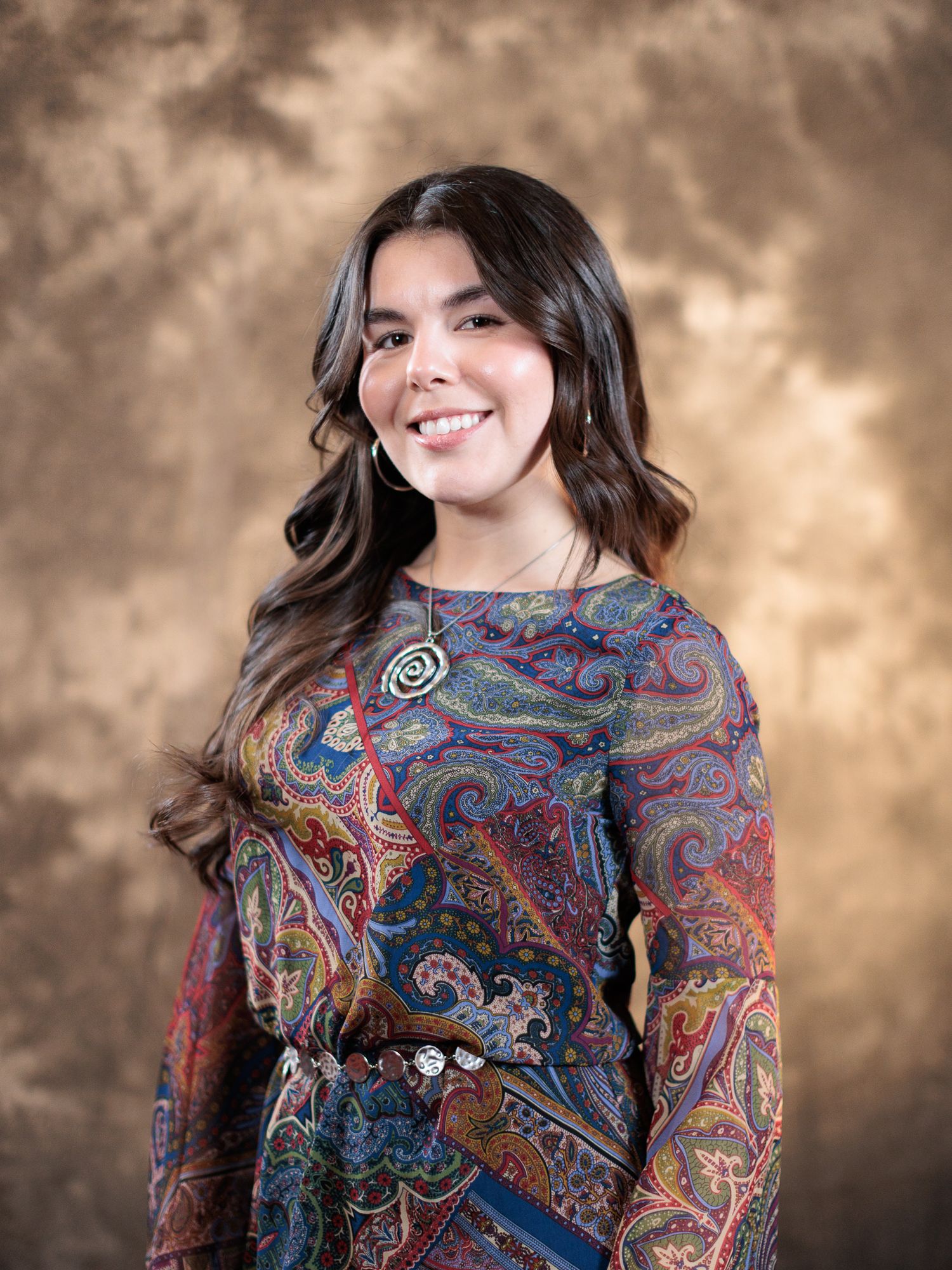 Young woman smiling in colorful paisley dress with spiral brooch against brown studio background.