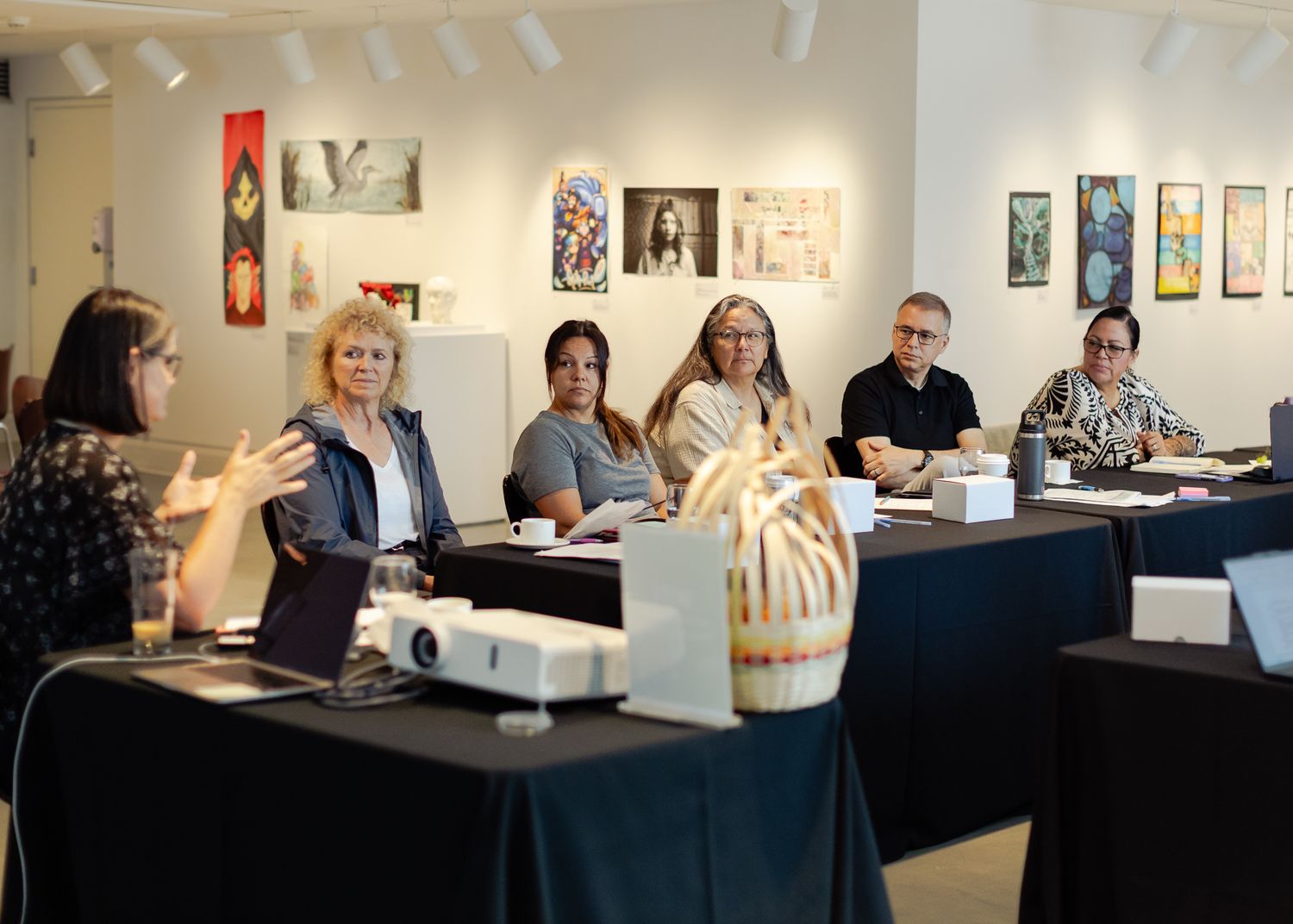 Group of people seated at black tables in an art gallery during a professional workshop or meeting.