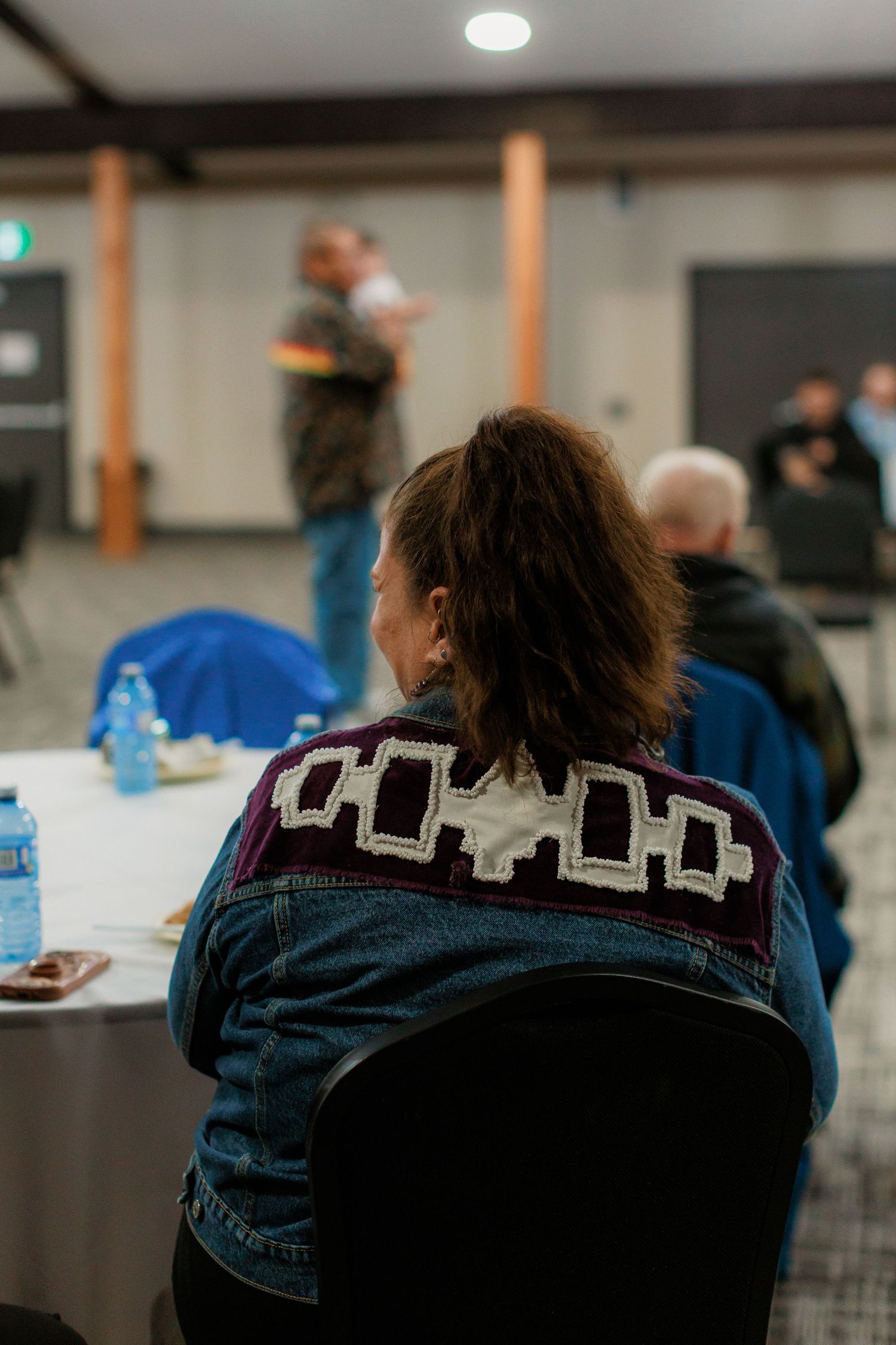 Woman in denim jacket with Indigenous beadwork pattern sits at a community meeting event.