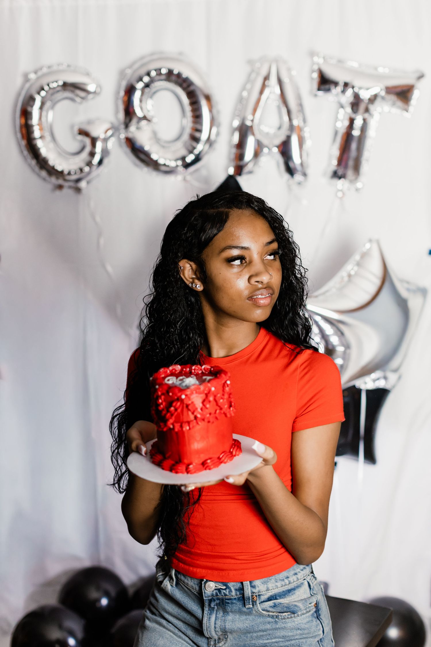 Young woman in red top holds red birthday cake in front of GOAT balloon letters and black balloons.