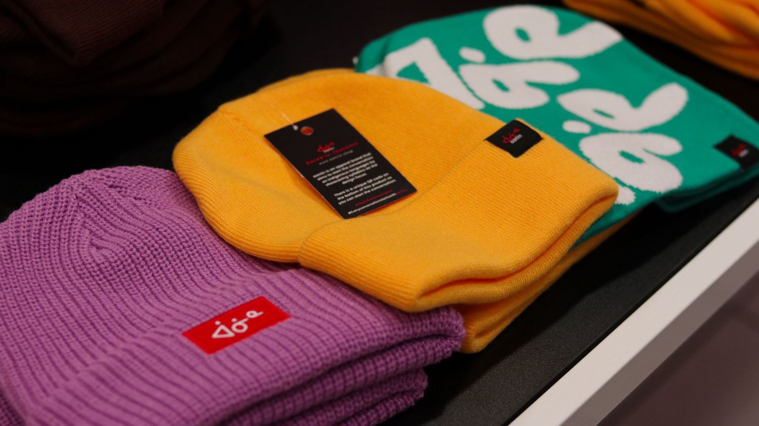 Colorful knit beanies in purple, yellow, and green with logo tags displayed on a retail shelf.