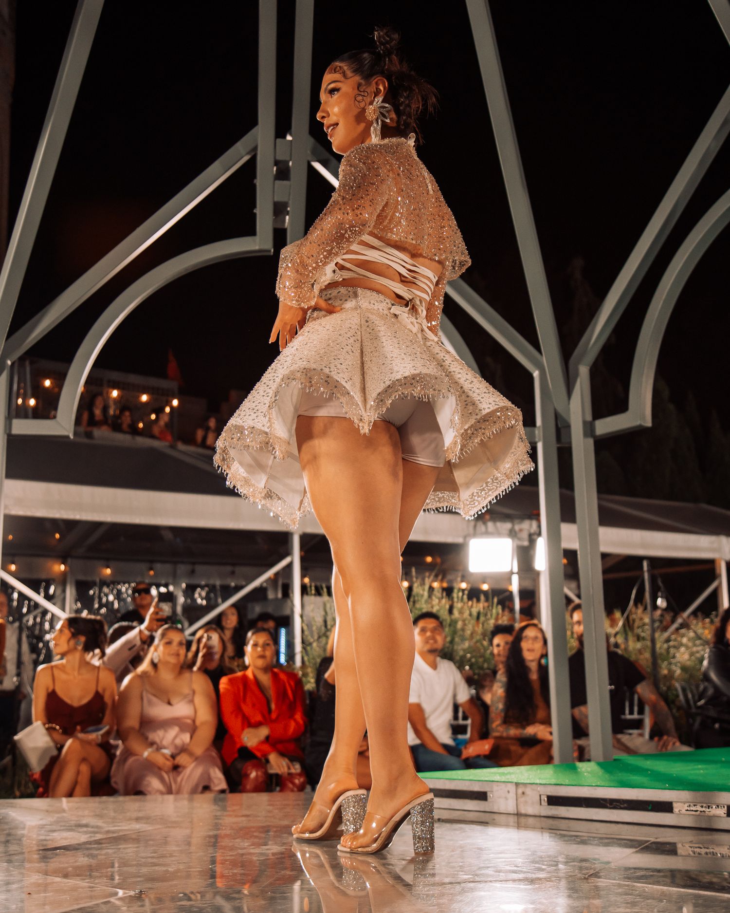 Model in sparkling white mini dress and heels walks runway before seated audience at night.