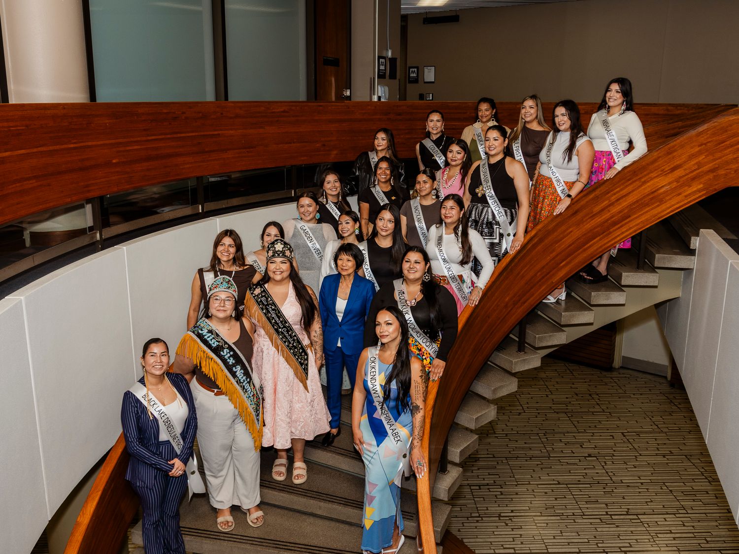 Miss Indigenous Canada pageant contestants pose on a curved staircase wearing sashes and formal wear.