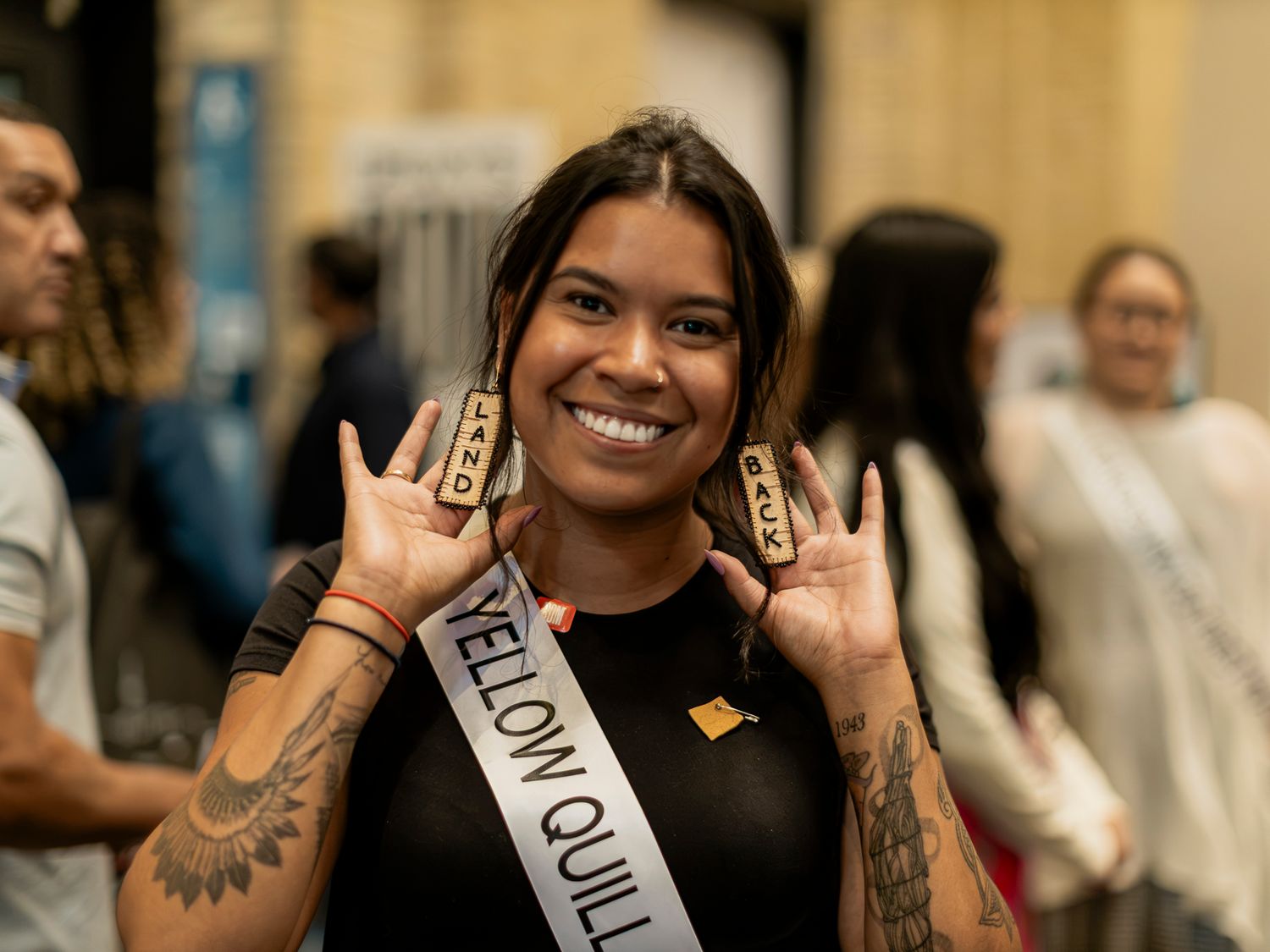 Smiling tattooed woman wearing a Yellow Quill sash holds up two small awards at an indoor event.
