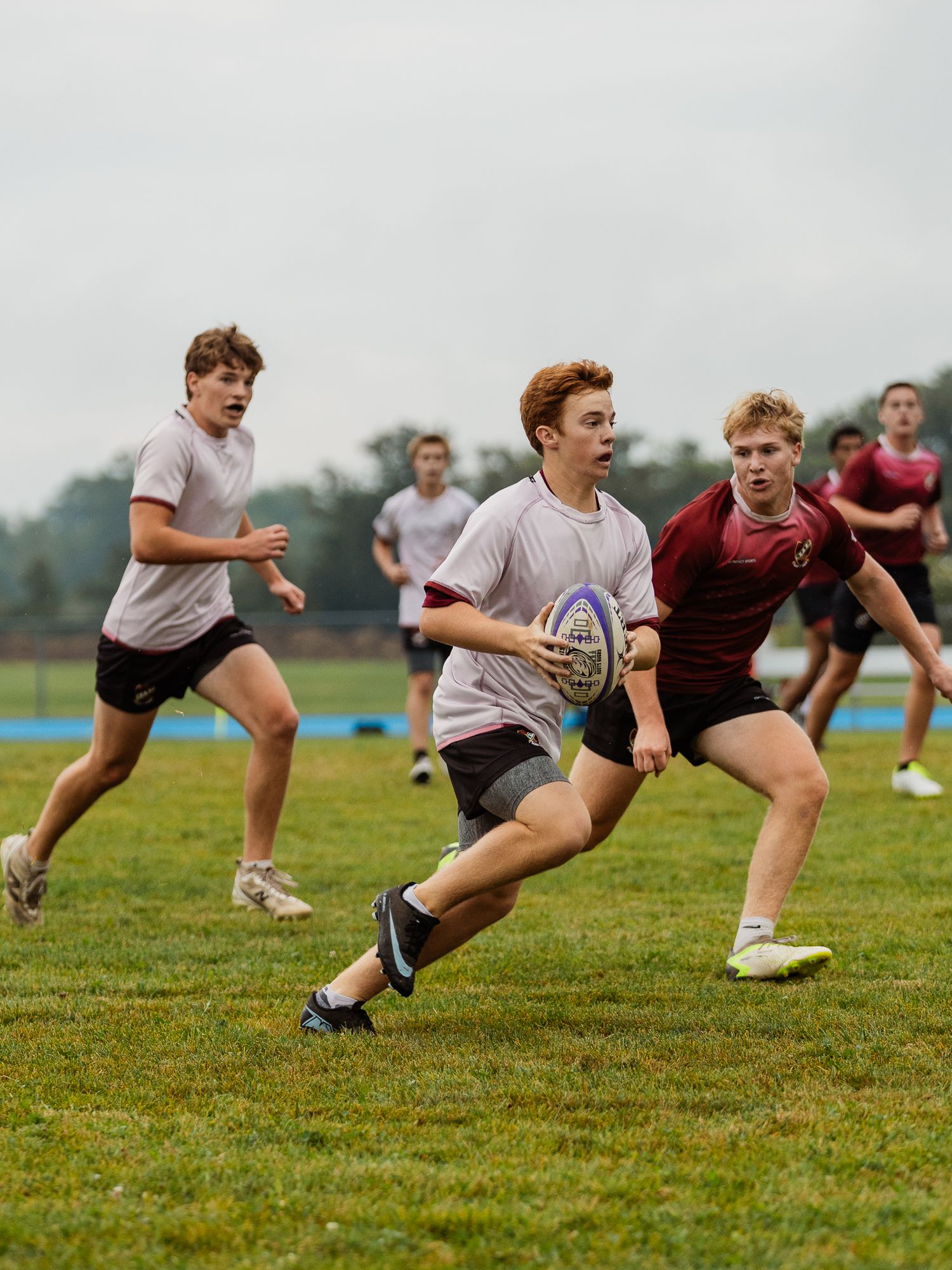 Teen rugby players compete on grass field, one carrying ball while defenders pursue during match.