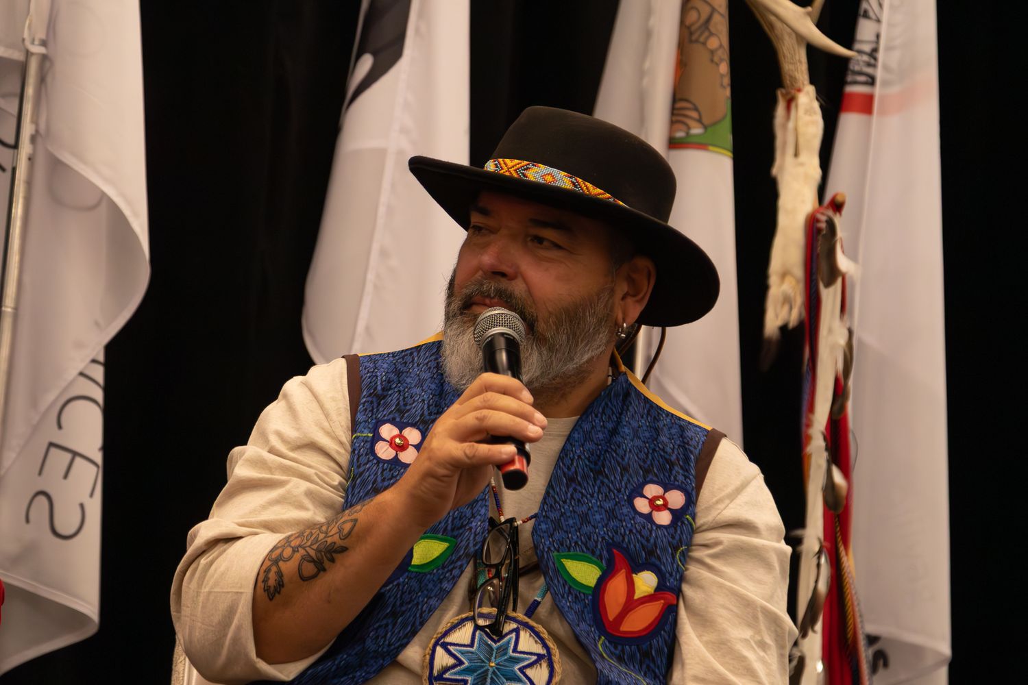 Native American man in black hat and beaded vest speaks into microphone at cultural event.