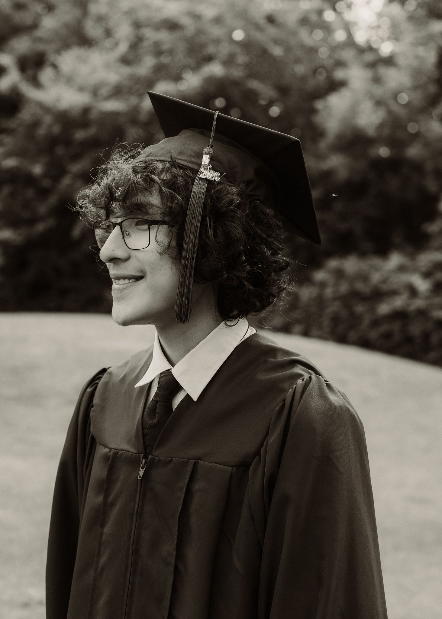 Smiling graduate in cap and gown poses outdoors in black and white portrait photo.