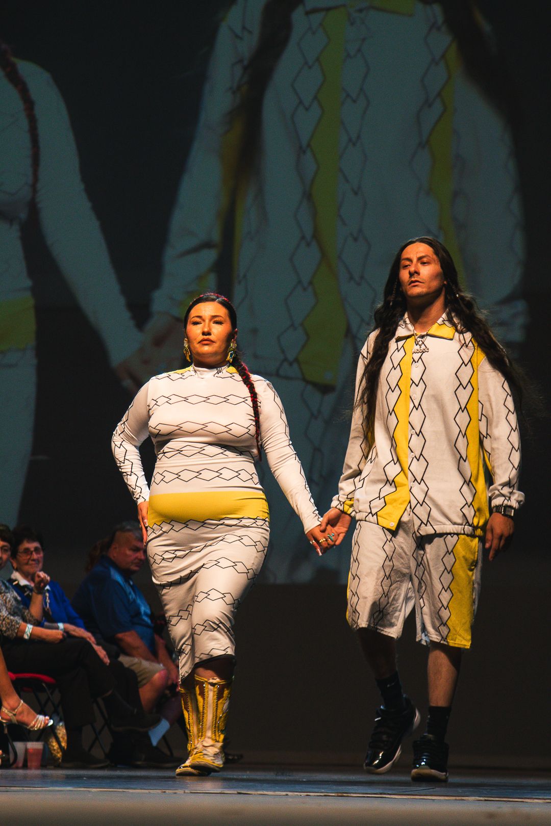 Two models walk a runway in white and yellow Indigenous-inspired fashion designs at a cultural fashion show.