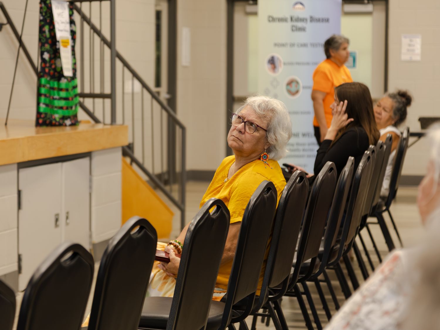 Elderly woman in yellow seated in row of black chairs at a chronic kidney disease clinic event.