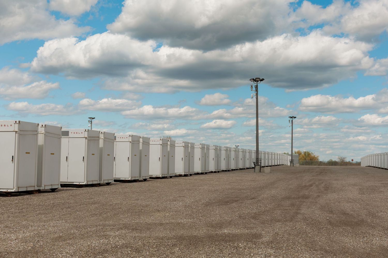 Row of white battery energy storage units at a large-scale grid facility under a partly cloudy sky.